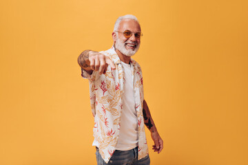Portrait of positive man in plant print shirt pointing to camera. Photo of handsome guy in bright summer clothes on orange background