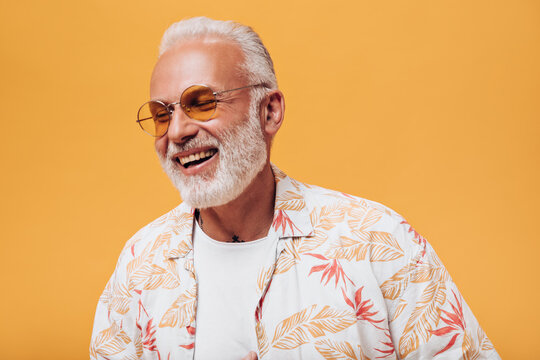 Awesome Man In Orange Eyeglasses Laughing On Isolated Background. Gray-haired Adult With Beard In Light Outfit Is Smiling Into Camera