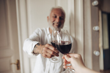 Handsome man in white outfit clinking glass with friend. Smiling bearded adult guy with tattoos on his hand with red wine