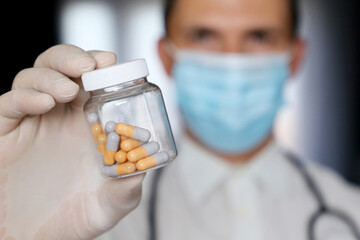 Doctor in medical mask holding bottle of pills, male hand in latex glove with medication in capsules close up. Concept of pharmacist, drugs, antibiotics or vitamins