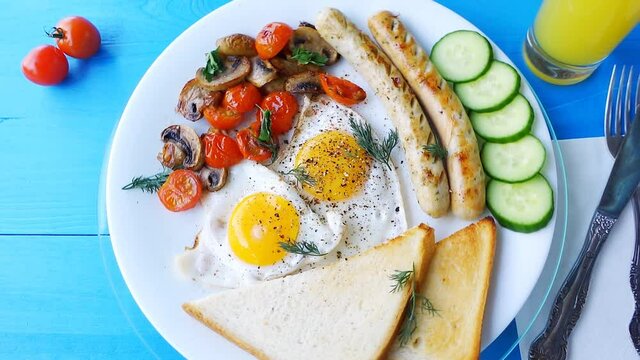 Slow motion loop video of a breakfast of fried eggs, mushrooms, tomatoes, sausages and bread, rotating on a white plate on a wooden blue table.	