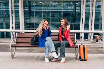 Two happy girls sitting on a bench near airport, with luggage. Air travel, summer holiday