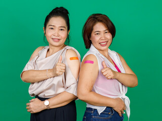 Two middle aged Asian women showing their arms with bandage patch showing they got vaccinated for Covid 19 virus on green background. Concept for Covid 19 vaccination