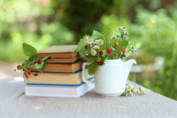 Red wild ripe strawberries and a cup with flowers and leaves from a bush of strawberries on a stack of books on a table in the garden