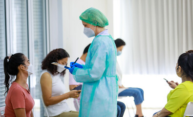Fototapeta premium Group of diverse citizens wear face mask in Coronavirus vaccinating queue line sitting checking his personal information from paper board in hands of full protection suit doctor