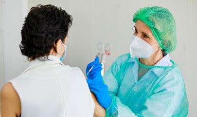 Caucasian senior female doctor in PPE full hazard protection uniform with face mask using syringe needle inject vaccine to woman patient shoulder at hospital vaccinating desk