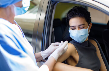 Fototapeta na wymiar Portrait closeup shot of female wearing face mask sitting in car receiving coronavirus vaccine from doctor wears hospital uniform using syringe and needle in drive through vaccination queue