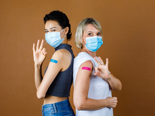 Caucasian female patients wears face mask standing look at camera lean on each other back showing okay or OK hand sign at colorful plaster together after vaccination in front brown background