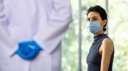 Female patient wears face mask sit wait frightened and fear while doctor in white lab coat and blue rubber gloves hold and hide vaccine syringe needle in hand behind back in blurred foreground