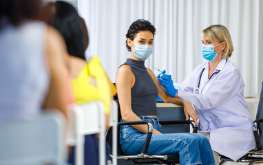 Obraz premium Cute female wears face mask sit look at camera while Caucasian woman doctor in white lab coat blue gloves and stethoscope injecting shot vaccine on shoulder when other patients wait in queue line