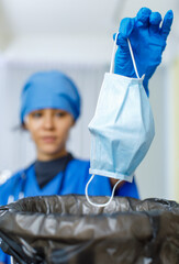 Closeup shot of used face mask was dumped into garbage bag trash can by female happy smiling doctor in blue hospital uniform rubber gloves and stethoscope in blurred background after pandemic ended