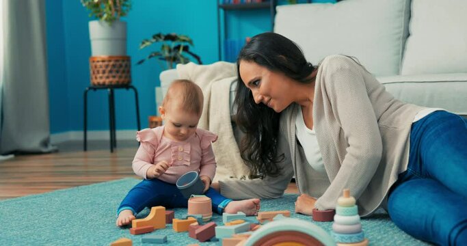 Funny, cute girl is playing with blocks with mother on carpet, she takes plastic container to her mouth, holds it, woman asks daughter to take it out, child lets go of toy and laughs