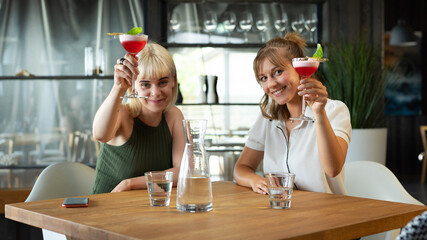 Two Female Friends Raising the Glass While Feeling Happy Together in a Modern Restaurant Sitting Behind a Wooden Table With Pink Cocktails and Glasses of Water.