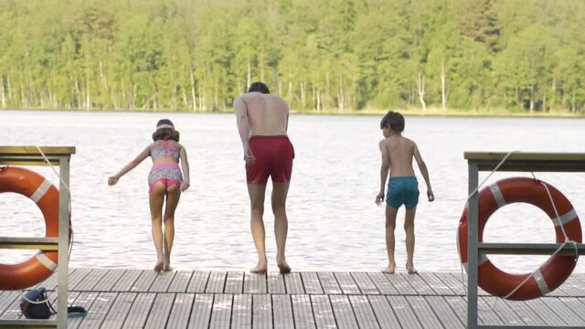 Happy family have vacations at the lake. Father with daughter and son jump from the pier together. Boy and girl diving from the dock into the water. Children having fun.