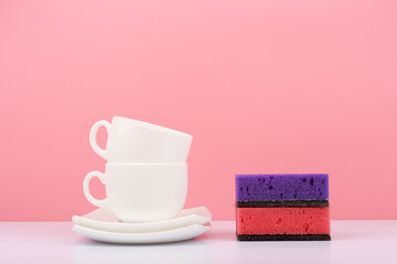 Dishwashing and housework concept. Clean white ceramic plates, cups and colorful cleaning sponges against bright pink background. 
