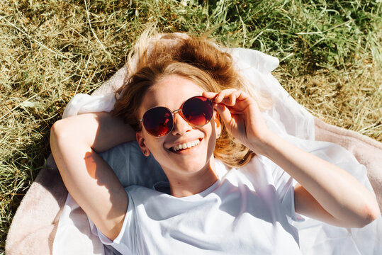 Top view portrait of laughing young woman with glasses lying on the lawn on sunny summer day. Happy beautiful girl resting on blanket on the grass and smiling at the camera, outdoors. Summer lifestyle