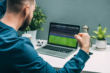 Over the shoulder view of euphoric young man watching soccer play live broadcast on his laptop and cheering for favourite team
