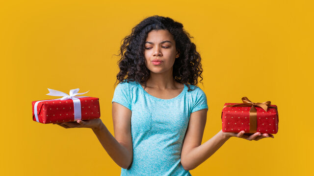 Pensive Dark Skinned Girl Holding Two Gift Boxes In Both Hands Deciding Which One To Choose