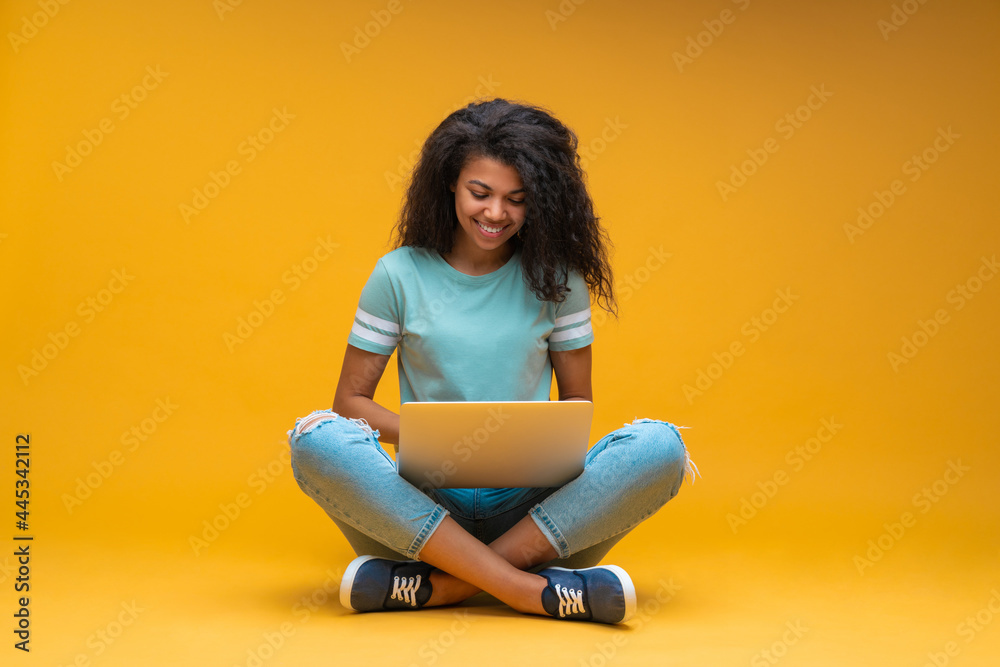 Wall mural beautiful young smiling dark skinned woman in casual outfit, sitting isolated on bright colored yell