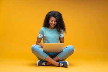 Beautiful young smiling dark skinned woman in casual outfit, sitting isolated on bright colored yellow background and working on her laptop