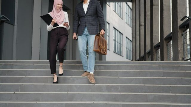 Full length of muslim woman and man walking together near modern office building an discussing some working moments. Two co workers with smartphones, clipboard and suitcase outdoors.