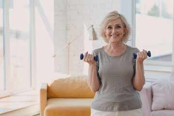 Elderly caucasian old aged woman portrait gray haired doing exercises with dumbbells in casual wear at home apartment living room