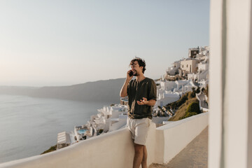 Fototapeta na wymiar Cheerful man talks on phone and smiles outside. Brunette guy in dark t-shirt and white shorts enjoys sea and city view.