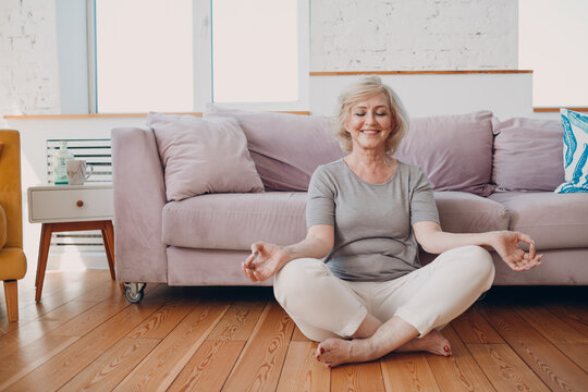 Senior Adult Smiling Woman Practicing Yoga At Home Living Room. Elderly Relaxed Female Sitting In Lotus Pose And Meditating Zen Like