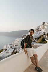 Fototapeta na wymiar Young brunette man in white shorts and dark-green t-shirt leans on white fence. Guy with backpack poses on background of city and sea.