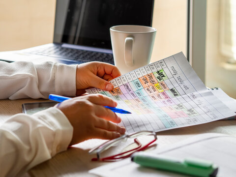 Woman Hands Holding A Schedule With Time Blocking Tasks For Productivity And Time Management. Work Desk With Laptop, Coffee Cup, Glasses And Colorful Weekly Planning. Remote Work From Home