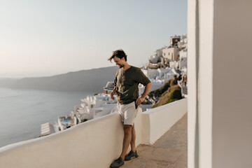 Fototapeta na wymiar Full-length portrait of brunette man in white shorts, dark green t-shirt looks at sea. Young happy guy smiles and enjoys city view.