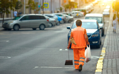 Municipal worker clean up street in the evening. worker in orange uniform collecting garbage from road and sidewalk. Woman in orange uniform with broomstick and scoop for garbage sweep city street