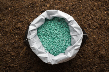 Opened plastic bag with green complex fertiliser granules on dark soil background. Closeup. Product...