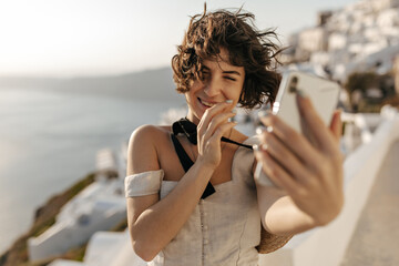 Curly short-haired woman in beige dress laughs and takes selfie in old Greek city. Happy brunette lady in stylish outfit holds phone and smiles on sea background.