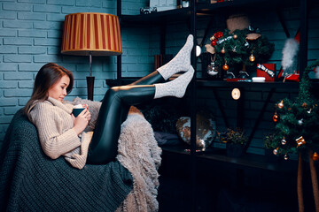 Fototapeta na wymiar Pretty woman with cup of cocoa sitting on chair. Christmas tree and decorations on shelf. New year's atmosphere. Warm cozy blankets on chair. Home loft interior with gray brick wall.