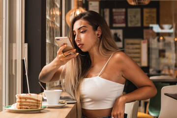 young blonde woman taking photo of a coffee and a cake in a coffee shop