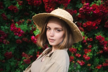 Elegant beautiful blonde young woman in a straw hat on the background of a bush of blooming roses.