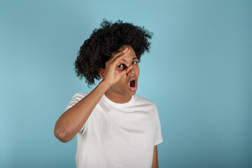 young african american man wearing basic white t-shirt on isolated pastel blue background peeking out in shock covering face and eyes with hand, looking through fingers with embarrassed expression.