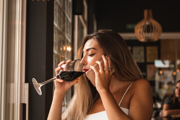 Young beautiful woman having a glass of red wine in a bar alone and talking on the cell phone