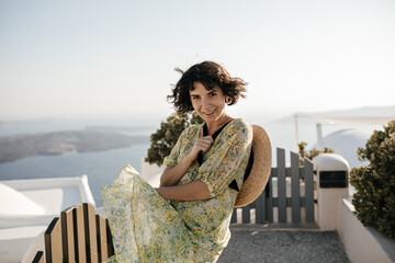 Fototapeta na wymiar Cheerful brunette woman in floral dress smiles sincerely outside. Happy lady in summer outfit holds boater and poses in Greek town with sea view.