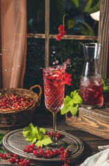 Glass and jug of fresh cold currant cocktail with splash and fly berries on the table near the window