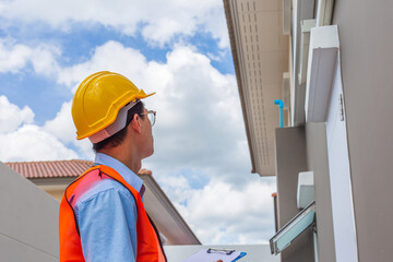 Asian inspector wearing a helmet and safety suit is taking notes after inspecting the structure and exterior of the house building