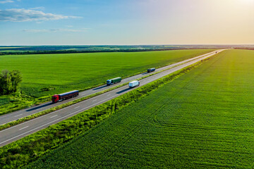 red green and blue trucks with container on the higthway sunset. cargo delivery driving on asphalt road along the green fields with goods. seen from the air. Aerial view landscape.convoys with cargo
