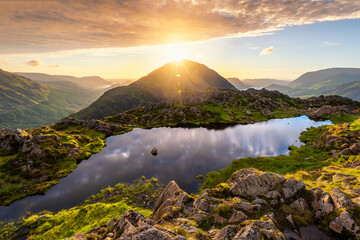 Beautiful majestic landscape with sun shining through mountain peak at Haystacks Tarn in the Lake District, UK. Breathtaking scenic viewpoint with calm reflections in body of water. - Powered by Adobe