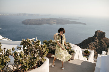 Happy curly woman in midi dress poses on terrace with sea view. Attractive brunette lady in floral...