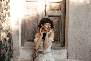 Brunette woman in beige dress with straw bag coquettishly poses outside. Curly short-haired lady sits near old house with wooden door.
