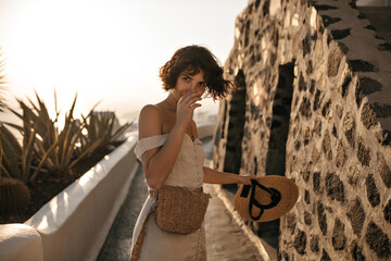 Brown-eyed brunette woman in beige dress with straw bag holds wide-brimmed hat and moves along path...