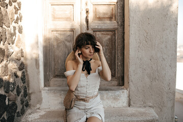 Curly brunette woman in beige dress with straw bag gently touches hair. Lady in stylish outfit poses outside and sits near old house with wooden door.