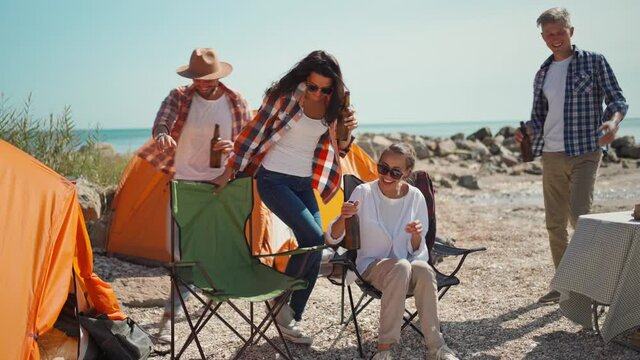 joyful friends on picnic returning from walks to camsite, playfuly sits at campchairs, drinking cold beer and laughing