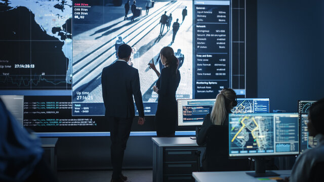 Male and Female Government Officers Discuss Working Matters next to a Computer and Big Digital Screen with Satellite Surveillance Footage with Face Recognition Software. Police Investigators at Work.